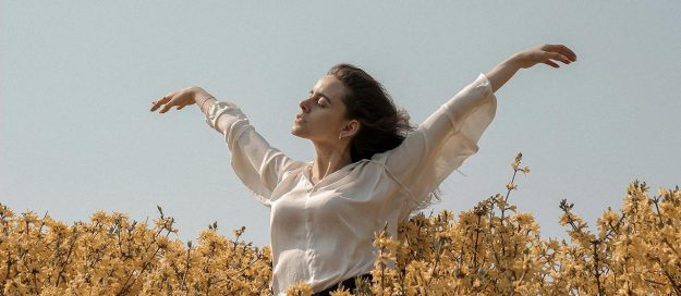 Mujer respirando relajada con los brazos abiertos rodeada de muchas flores blancas y amarillas