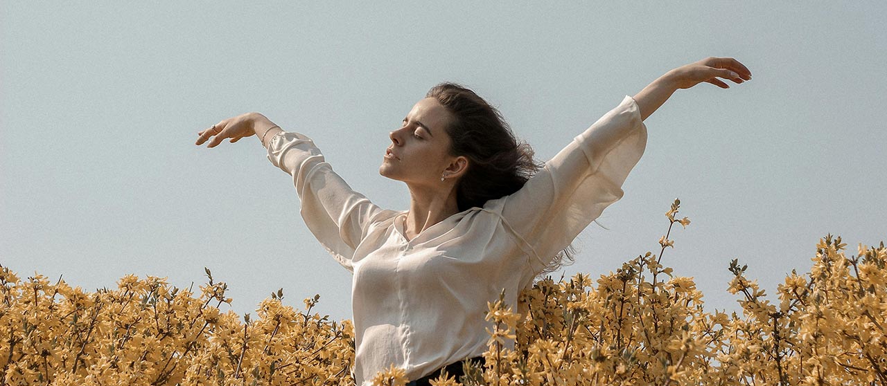 Mujer respirando relajada con los brazos abiertos rodeada de muchas flores blancas y amarillas